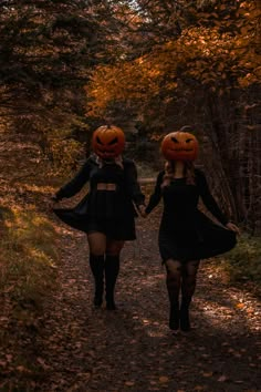 two women wearing pumpkin heads walk down a path in the woods with their hands on each other's backs