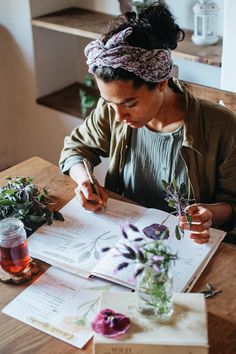 a woman sitting at a table with flowers and writing in a book on top of it