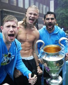 three shirtless men pose with a trophy in the middle of a street while another man holds his mouth open