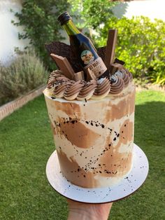 a hand holding a cake with chocolate frosting and wine bottle on top in the grass