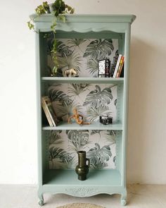 a green bookcase with plants and books on it in front of a wallpapered wall