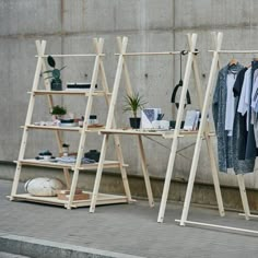 two wooden racks with clothes hanging on them next to a concrete wall and potted plants