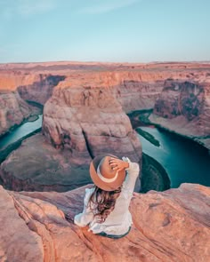 a woman wearing a hat sitting on top of a cliff next to a body of water