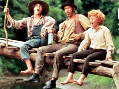 three people sitting on top of a wooden bench in front of a river and trees