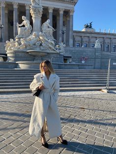 a woman standing in front of a building wearing a white coat and black heels with her hand on her hip