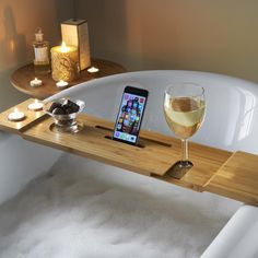 a cell phone is sitting on a wooden tray in a bathtub next to a glass of wine