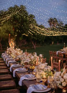 an outdoor dinner table set up with white flowers and greenery, lit by string lights