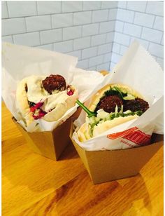 two take out sandwiches with meatballs and vegetables in paper wrappers on a wooden table