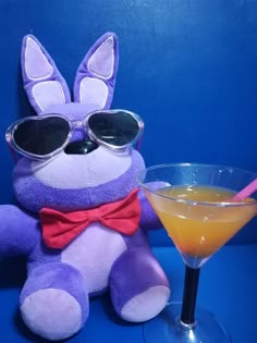 a purple stuffed animal sitting next to a glass filled with orange juice and wearing sunglasses