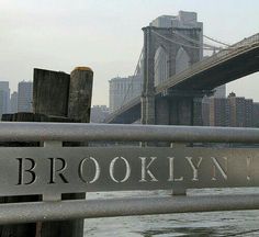 there is a sign that says brooklyn in front of the water and a bridge behind it