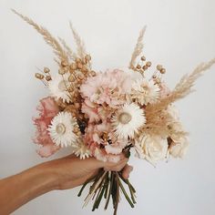 a hand holding a bouquet of flowers against a white wall with dry grass in the foreground