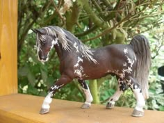 a toy horse standing on top of a wooden table next to a tree and bushes