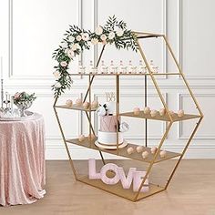 a gold hexagonal shelf with candles and flowers on it next to a pink table cloth