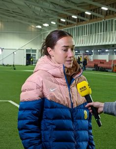 a woman in a blue and pink jacket holding a microphone while standing on a soccer field