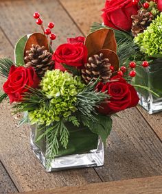 two glass vases filled with red roses and greenery on a wooden table next to each other