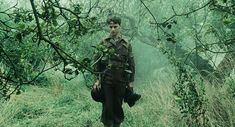 a man standing in the middle of a lush green forest holding a bag and looking at the camera