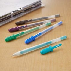 five pens are lined up on a table next to a notebook and pencils with erasers