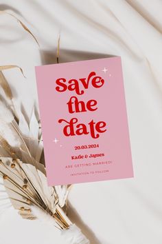 a pink save the date card sitting on top of a white sheet with dried grass