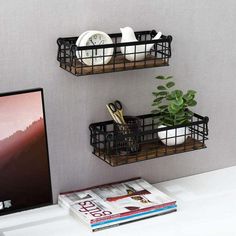 two black metal baskets on the wall above a computer desk with books, magazines and plants