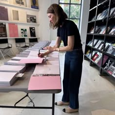 a woman standing at a table with several books on it and writing in a notebook