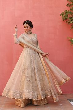 a woman in a white and gold lehenga standing against a pink wall with her hands up