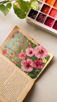 an open book sitting on top of a table next to flowers and watercolors