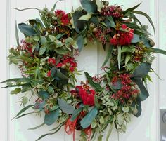 a wreath with red flowers and greenery hangs on a door handle, in front of a white door