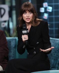 a woman sitting on top of a blue couch holding a microphone in her hand and wearing a black suit