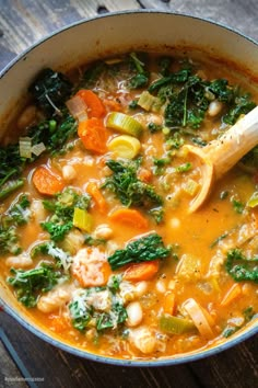 a pot filled with soup and vegetables on top of a wooden table next to a spoon