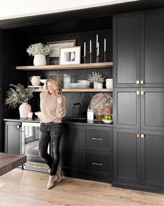 a woman standing in front of a kitchen counter with black cabinets and white vases