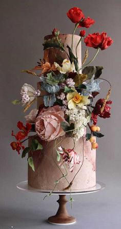 a three tiered cake decorated with flowers and greenery on a wooden stand in front of a gray background