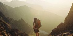 two people standing on the side of a mountain with backpacks over their heads looking at the valley below