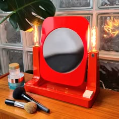 a red vanity mirror sitting on top of a wooden table next to makeup brushes and a potted plant