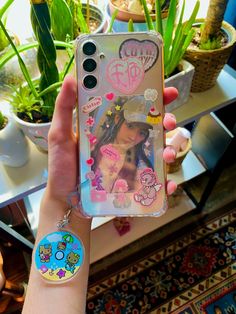 a person holding up a cell phone case with stickers on it and plants in the background