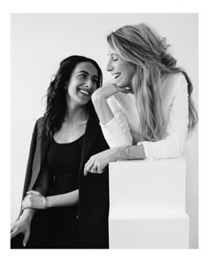 two women sitting next to each other on a white surface with one woman leaning against the wall