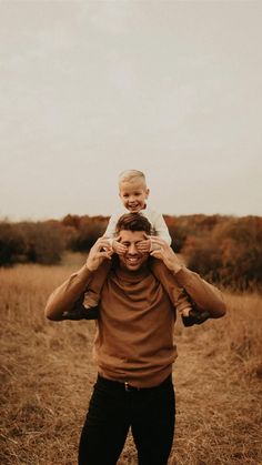 a man holding a child on his shoulders in a field with dry grass and trees