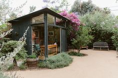 a small building with lots of plants and flowers in it's front yard, next to a bench