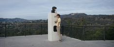 a person standing next to a sculpture on top of a hill with mountains in the background