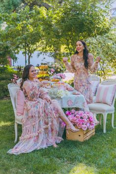 two women sitting at a table with pink flowers in the grass and one woman standing next to her