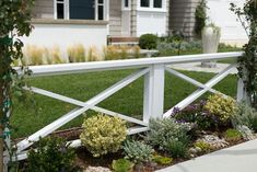 a white wooden fence in front of a house with green grass and flowers around it