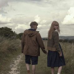 a man and woman walking down a dirt road holding hands with each other on a cloudy day