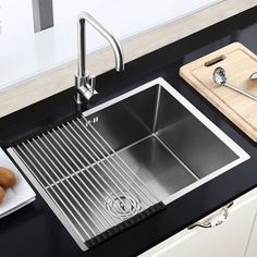 stainless steel kitchen sink with grid strainer and cutting board on black countertop next to utensils