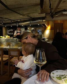 a woman holding a baby in her arms while sitting at a table with food and wine glasses