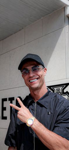 a man in black shirt and hat giving the peace sign with his hand while standing next to a building