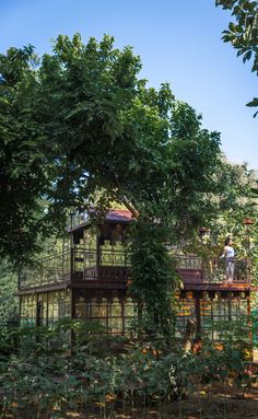 the house is surrounded by many trees and plants in front of it, with two people standing on the balcony