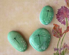 three green rocks with dragonflies painted on them and flowers in the backgro