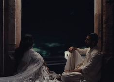 a man and woman sitting on the ground in front of an archway, dressed in white