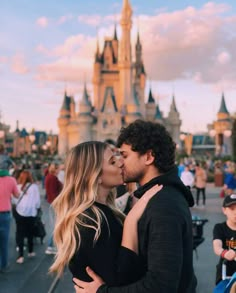 a man and woman kissing in front of a castle at disneyland world with people walking around