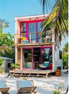 a pink and purple house on the beach with chairs around it, in front of palm trees