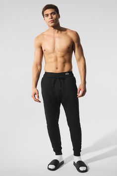 a shirtless man in black pants and slippers stands against a white background with his hands on his hips